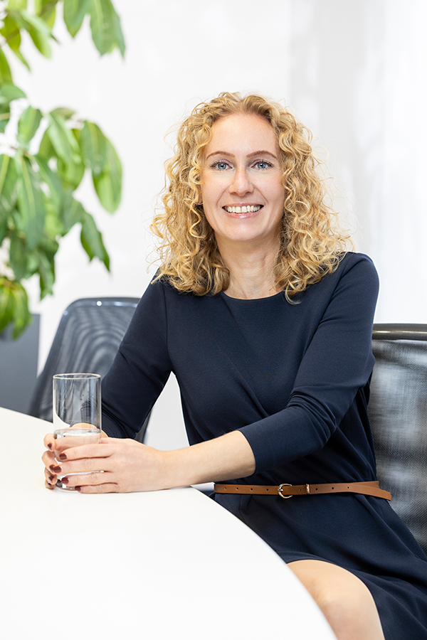 Finanzbuchhalter bei der MIZ. Frau mit lockigem Haar am Tisch mit Glas Wasser lächelnd.