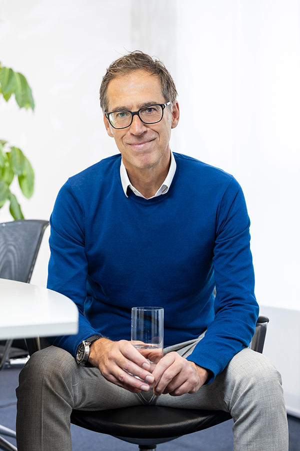 Rechtsberatung bei der MIZ. Mann in blauem Pullover hält ein Glas, sitzt lächelnd im Büro.