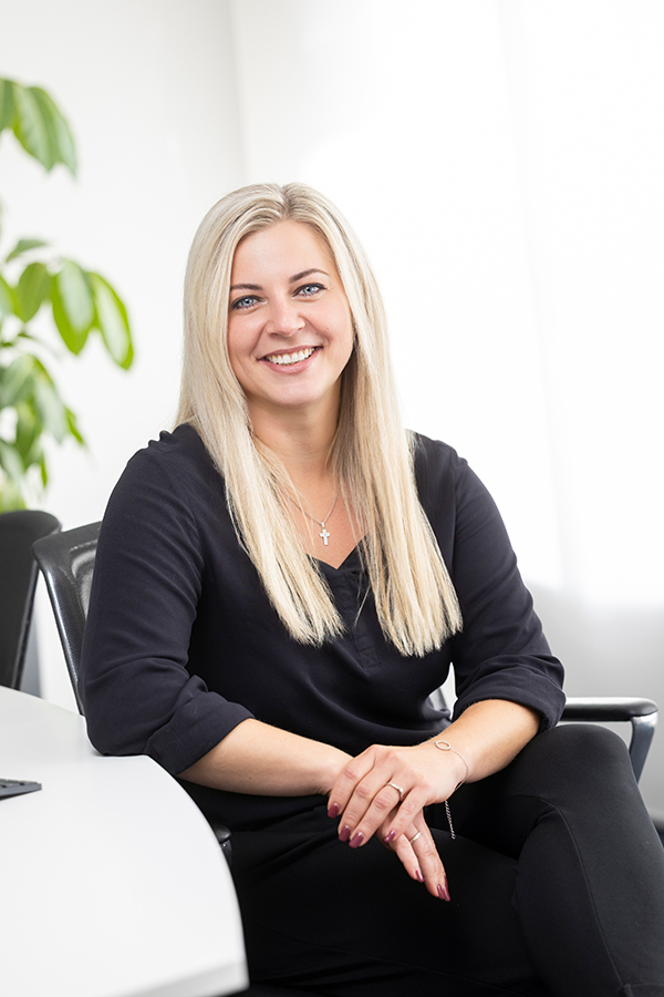 Steuerassistent bei MIZ. Lächelnde Frau im schwarzen Outfit sitzt an einem Tisch.