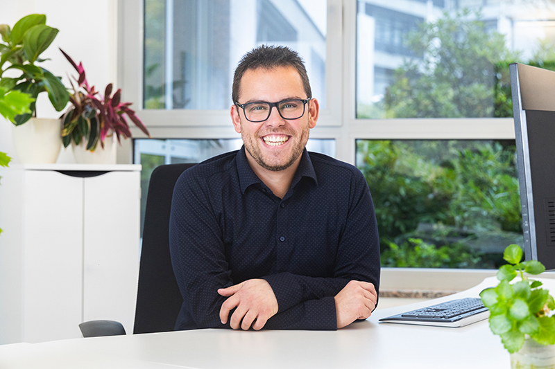 Steuerfachangestellter bei MIZ. Mann mit Brille lächelt am Schreibtisch in einem modernen Büro.